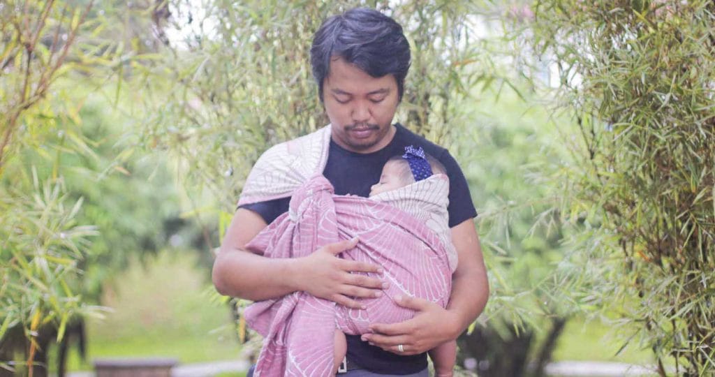 dad stands with trees in background and new baby in babywearing wrap