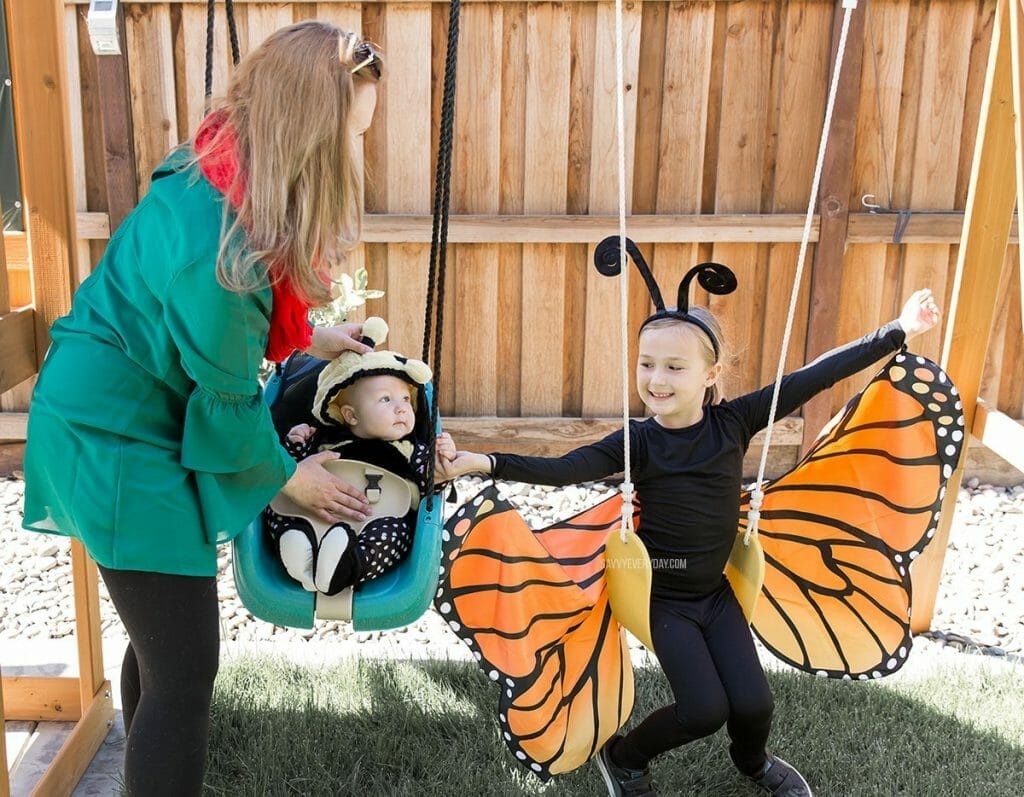 helping baby in a bee costume on a swing set while girl in butterfly costume sits on a swing with outstretched wings