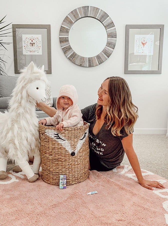 baby stands in a wicker basket wearing hooded sweater next to mom and giant stuffed llama in pink and white nursery room