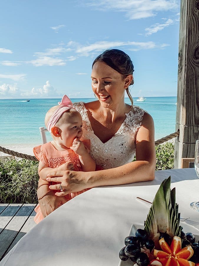 mom in wedding dress smiles down at baby in pink dress sitting on her lap and teething on her fingers