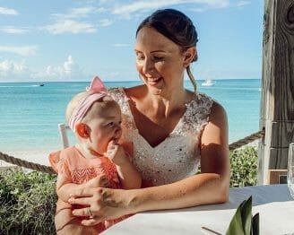 mom in wedding dress smiles down at baby in pink dress sitting on her lap and teething on her fingers