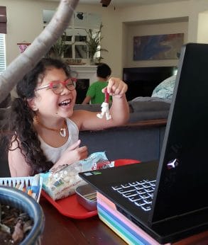 girl with glasses smiles as she holds up her project to friends in camp zoom classroom on her computer