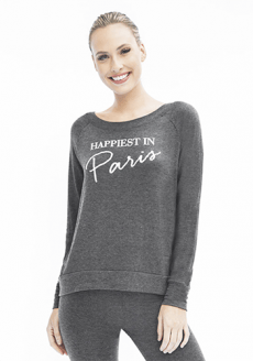 White woman with blonde hair smiling at camera wearing grey leggings and grey long sleeve shirt that says "Happiest in Paris"