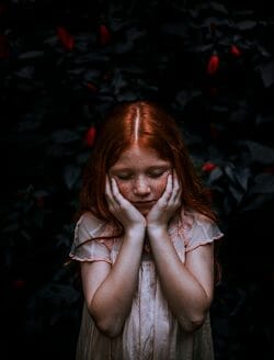 redhaired girl with hands over her cheeks in front of dark background- looks worried