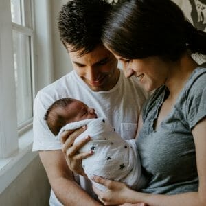new postpartum parents hold swaddled newborn in their arms and smile