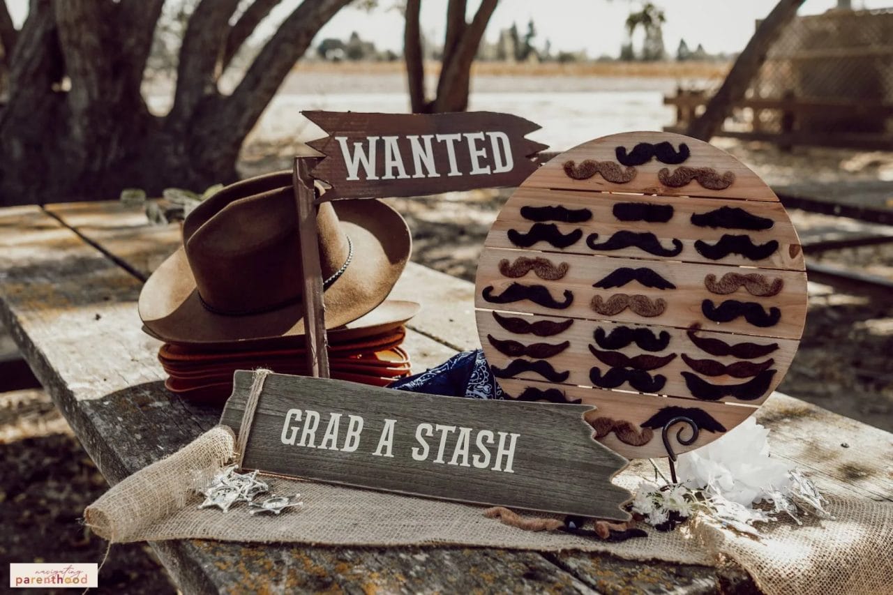 Table with mustaches taped to a round wooden board with a sign that says "grab a stash" and stacked cowboy hats at the Wild West first birthday party